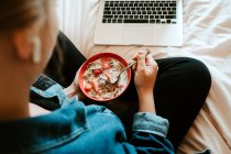 From above female with earphones eating cereals with strawberries for breakfast in a bowl while sitting with legs crossed on soft bed and using computer in cozy apartment — Stock Photo