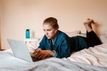 Mujer pensativa enfocada en verdaderos auriculares inalámbricos y ropa casual acostada en la cama usando un portátil contra la pared beige en un apartamento ligero - foto de stock