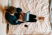 From above unrecognizable barefoot female in casual clothes focusing on screen and working on laptop while sitting on soft bed with smartphone and wireless earphones in cozy contemporary apartment — Stock Photo