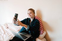 Joven mujer feliz en los auriculares inalámbricos y chaqueta de mezclilla utilizando el teléfono inteligente para tomar una selfie y un ordenador portátil mientras se relaja en la cama en el apartamento moderno - foto de stock