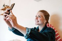 Радісна молода жінка в бездротових навушниках і джинсовій куртці посміхається на екрані і приймає селфі зі смартфоном, розслабляючись на ліжку в сучасній квартирі — стокове фото