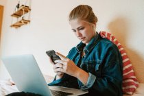 Jovem fêmea em fones de ouvido sem fio e jaqueta jeans usando smartphone e laptop enquanto relaxa na cama no apartamento moderno — Fotografia de Stock