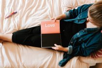 Von oben eine zierliche Frau in schwarzer Freizeitkleidung und Jeansjacke, die ein geöffnetes Tagebuch mit schwarzen und rosa Seiten in der Hand hält, während sie allein auf einem weichen Bett zu Hause ruht — Stockfoto