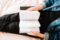 From above crop curious woman in black clothes and denim jacket flipping book with interest preparing to read while chilling alone on soft bed at home — Stock Photo