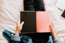 Von oben sieht man eine unkenntliche Frau in schwarzer Freizeitkleidung und Jeansjacke, die ein geöffnetes Tagebuch mit schwarzen und rosafarbenen Seiten in der Hand hält, während sie allein auf einem weichen Bett zu Hause liegt. — Stockfoto