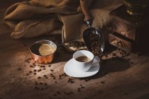 Свіжа чорна кава в білій керамічній чашці покладена на блюдце біля кавоварки та кавових зерен на дерев'яному столі — стокове фото
