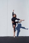 Повне тіло жіночої танцівниці в чорному костюмі та взутті, що виконує поставу із закритими очима, стоячи на кінчику на одній нозі на білій стіні з падаючою тіні — стокове фото
