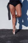Bailarina femenina irreconocible recortada en traje negro y zapatos puntiagudos realizando postura de puntillas mientras está de pie contra la pared blanca con sombra que cae - foto de stock