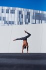Back view of unrecognizable shirtless sportsman performing handstand with one hand while doing exercises against wall and blue sky on rooftop of modern building — Stock Photo
