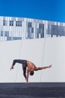 Вид ззаду невпізнаваного спортсмена без сорочки, який виконує сумочку однією рукою під час виконання вправ проти стіни та блакитного неба на даху сучасної будівлі — стокове фото