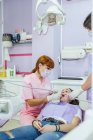 Female dentist in uniform and mask curing patient teeth with female assistant in modern dental clinic — Stock Photo