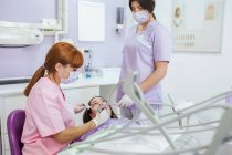 Dentista feminina de uniforme e máscara curando os dentes do paciente com assistente feminino na clínica odontológica moderna — Fotografia de Stock
