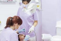 Dentista femenina en uniforme y mascarilla curando dientes de paciente con asistente femenina en clínica dental moderna - foto de stock