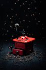 Vintage metal red coffee grinder placed on black wooden surface and falling coffee grains in dark room on black background — Stock Photo