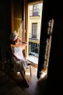 Serena hembra relajada en toalla blanca sobre cuerpo y cabeza sentada en silla junto a la puerta del balcón en apartamento con suelo de madera en día soleado y mirando hacia otro lado - foto de stock