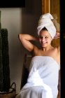 Happy female in white towel on body and head sitting on chair by the door leaned on hands in apartment on sunny day and looking away — Stock Photo