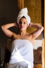 Donna felice in asciugamano bianco sul corpo e la testa seduta sulla sedia dalla porta appoggiata sulle mani in appartamento nella giornata di sole e guardando altrove — Foto stock