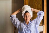 Улыбающаяся женщина с белым махровым полотенцем на голове, стоящая опираясь на руку и глядя на камеру в квартире в солнечный день — стоковое фото