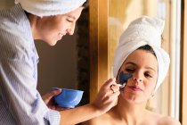 Cheerful female applying blue clay mask to face of serene girlfriend looking away with towel wrapped in head after bath while sitting near balcony in sunlight — Stock Photo