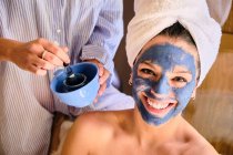 Anonymous female applying blue clay mask to face of cheerful woman looking at camera in white towel during procedure at home — Stock Photo