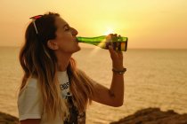 Smiling young lady with bottle of beer during sunset on seashore — Stock Photo