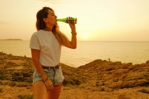 Улыбающаяся юная леди с бутылкой пива во время заката на берегу моря — стоковое фото