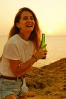 Jovencita sonriente con botella de cerveza al atardecer en la orilla del mar - foto de stock
