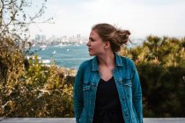 Jeune touriste féminine pensive veste en jean décontracté et robe noire debout près des arbres verts contre une vue imprenable sur la mer bleue et le ciel nuageux à Istanbul et détournant les yeux — Photo de stock