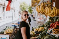 Vista laterale della donna positiva in abbigliamento casual e occhiali da sole guardando la fotocamera in piedi vicino al bancone della frutta nel mercato turco ed esplorando le merci mentre si cammina per le strade della città — Foto stock