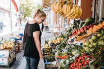 Vue latérale de la femme positive en tenue décontractée et lunettes de soleil debout près du comptoir de fruits dans le marché turc et explorer les marchandises tout en marchant dans les rues de la ville — Photo de stock