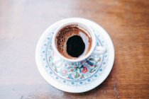 Сверху ароматный черный горячий турецкий кофе в хрупкой традиционной белой чашке и блюдце окрашены восточным узором стоя на деревянном столе — стоковое фото