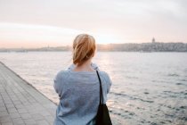 Visão traseira de viajante feminino anônimo em traje casual em pé no dique e admirando a vista do pôr do sol incrível colorido — Fotografia de Stock