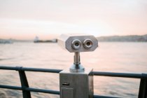 Amazing landscape of gray steel binocular locating on observation platform near black fencing against background of calm sea water reflecting and marvelous light pink sunset sky — Stock Photo