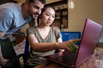 Baixo ângulo de feliz namorado ajudando asiático namorada trabalhando no computador em casa — Fotografia de Stock