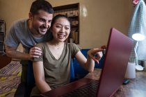 Faible angle de petit ami heureux aidant petite amie asiatique travaillant sur l'ordinateur à la maison — Photo de stock