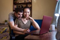 Faible angle de petit ami heureux aidant petite amie asiatique travaillant sur l'ordinateur à la maison — Photo de stock