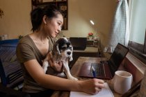 Visão lateral de freelancer feminino focado sentado na cadeira tomando notas no bloco de notas enquanto segura um cão trabalhando remotamente de casa — Fotografia de Stock
