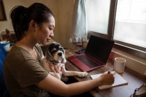 Вид сбоку сосредоточенной женщины-фрилансера, сидящей на стуле и делающей заметки на блокноте, держа в руках собаку, работающую дистанционно от дома — стоковое фото