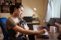 Side view of focused female freelancer sitting on chair taking notes on notepad while holding a dog working remotely from home — Stock Photo