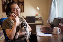 Freelancer femenina seria hablando en smartphone sobre un proyecto sentado en una silla trabajando remotamente en una computadora y bloc de notas mientras sostiene a un perro - foto de stock
