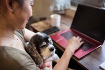 Зверху обрізаний веселий жіночий фрілансер, який працює дистанційно на ноутбуці, сидить на стільці, тримаючи собаку — стокове фото