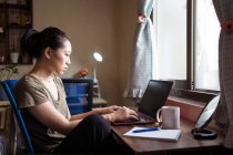 Seitenansicht einer asiatischen Freiberuflerin in lässigem T-Shirt und Brille, die am Tisch sitzt und am Computer surft, während sie zu Hause an einem Projekt online arbeitet — Stockfoto