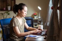 Vista lateral de mujer freelancer asiática en camiseta casual y anteojos sentados en la mesa y la computadora de navegación mientras se trabaja en el proyecto en línea en casa - foto de stock