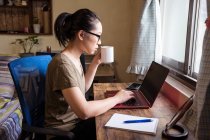 Side view of Asian female freelancer in casual t shirt and eyeglasses sitting at table and browsing computer while working on project online at home and drinking hot beverage from a mug — Stock Photo