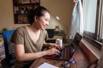 Vista lateral de mujer freelancer asiática en camiseta casual y anteojos sentados en la mesa y la computadora de navegación mientras se trabaja en el proyecto en línea en casa - foto de stock