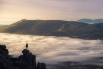 Remote unrecognizable hiker standing on edge of rock above cloudy foggy valley and admiring magnificent mountain landscape in morning — Stock Photo