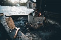 Brennendes Lagerfeuer und Baumstamm mit Axt in der Nähe einer Holzfällerhütte im verschneiten Wald an einem Wintertag in der Landschaft Finnlands — Stockfoto