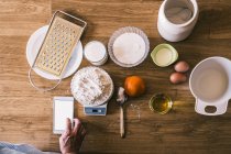 Top view of crop anonymous female browsing recipe on smartphone and weighing wheat flour on electronic scales while preparing ingredients for homemade pastry in kitchen — Stock Photo