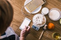 Top view of crop anonymous female browsing recipe on smartphone and weighing wheat flour on electronic scales while preparing ingredients for homemade pastry in kitchen — Stock Photo
