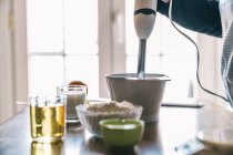 Vista laterale della casalinga anonima del raccolto utilizzando mixer durante la preparazione di pasta per muffin dolci al bancone della cucina con ingredienti per la ricetta — Foto stock